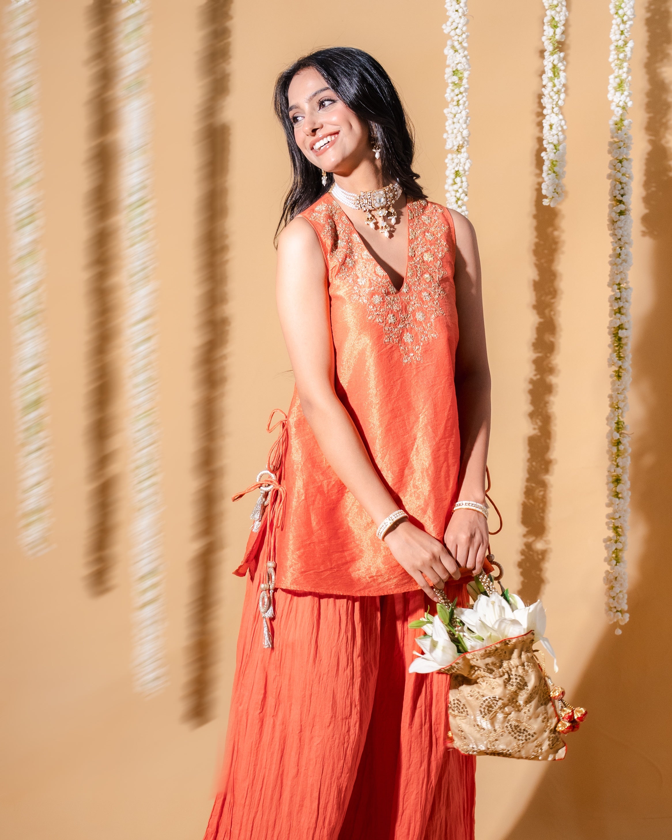 Woman in an orange rust outfit holding a basket bouquet against a beige wall with floral decorations.